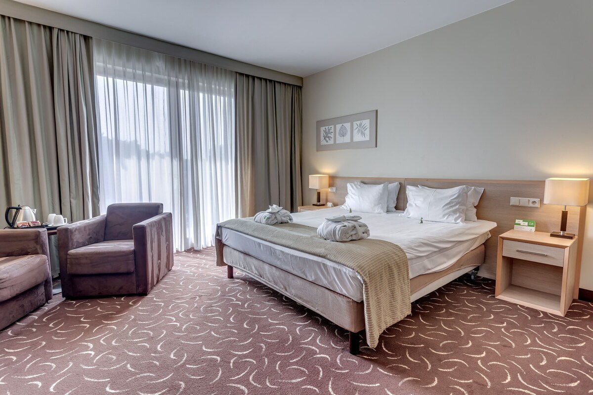 A hotel room with a large bed, two bedside tables with lamps, a chair, and a large window with curtains.