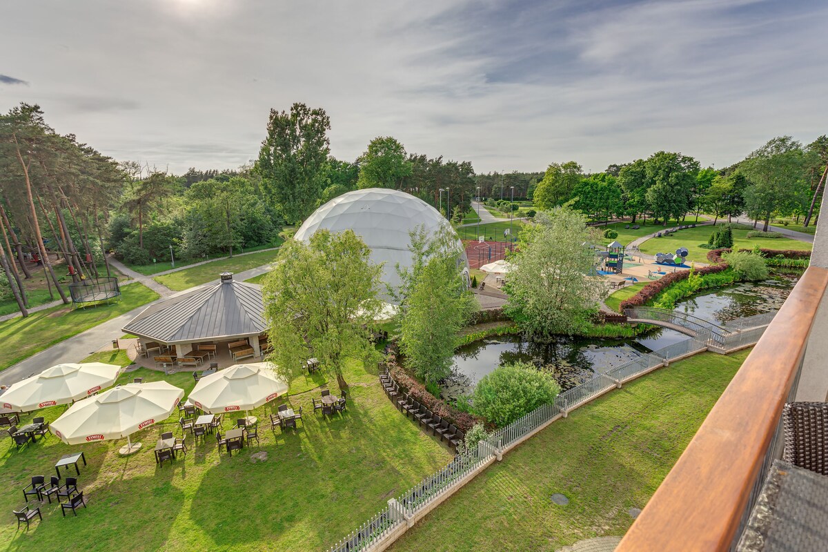 A view of a park with a large dome structure, outdoor seating, and a pond.