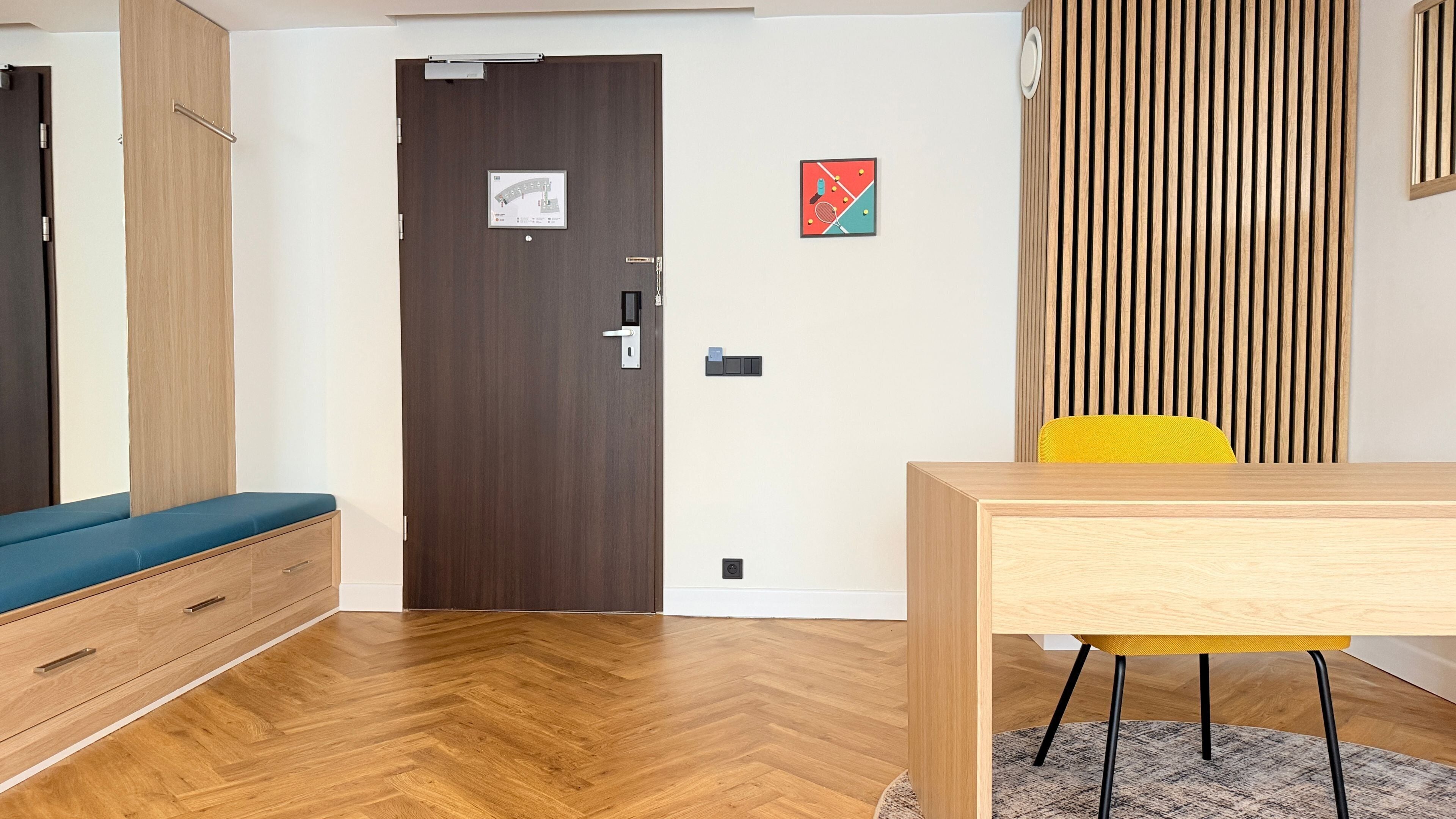A room with a wooden floor, a blue sofa, a wooden table with a yellow chair, a door with a lock, and a wall-mounted light fixture.