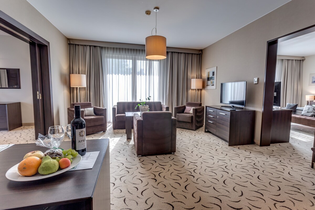 A hotel room with a dining area, a sofa set, a TV, and a large window with curtains.