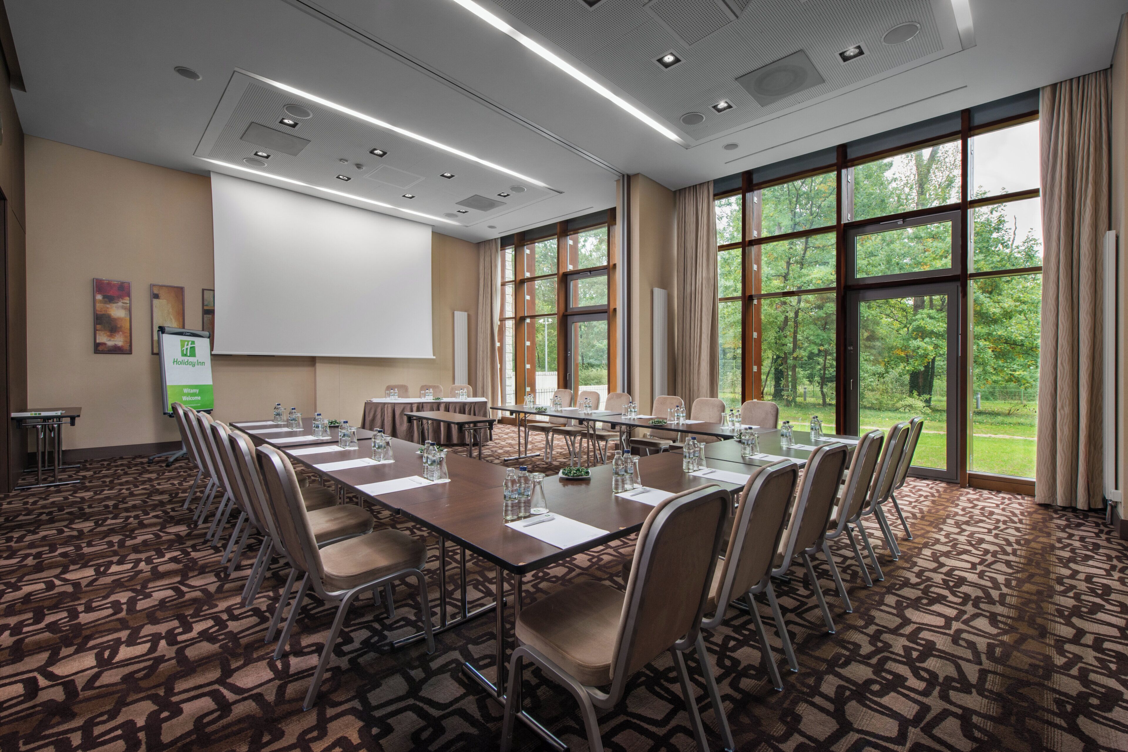 A conference room with a long table, chairs, a projector screen, and large windows with a view of greenery.