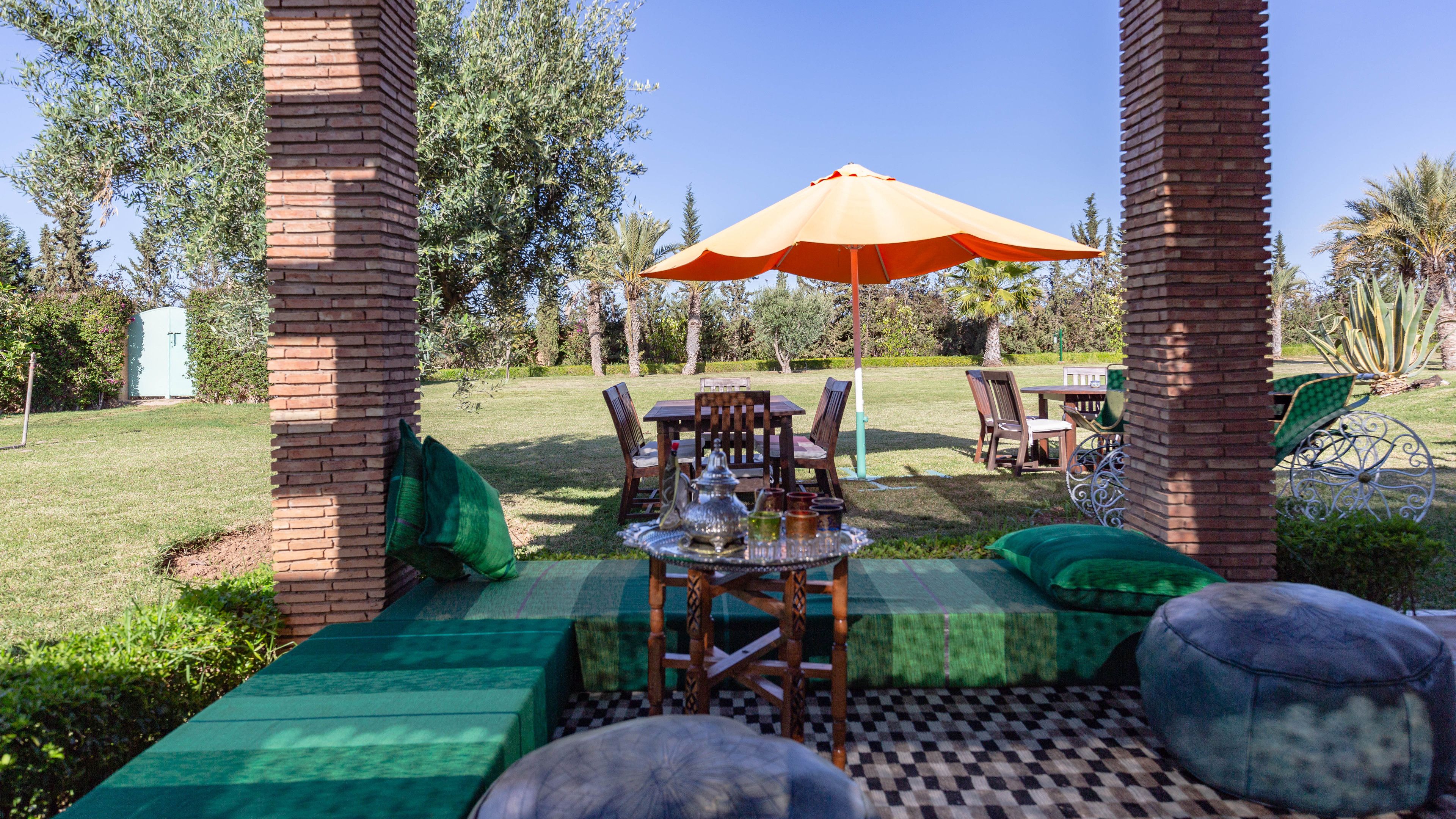 Un patio al aire libre con una mesa lista para comer, rodeado de pilares de ladrillo, cojines verdes y un suelo a cuadros.