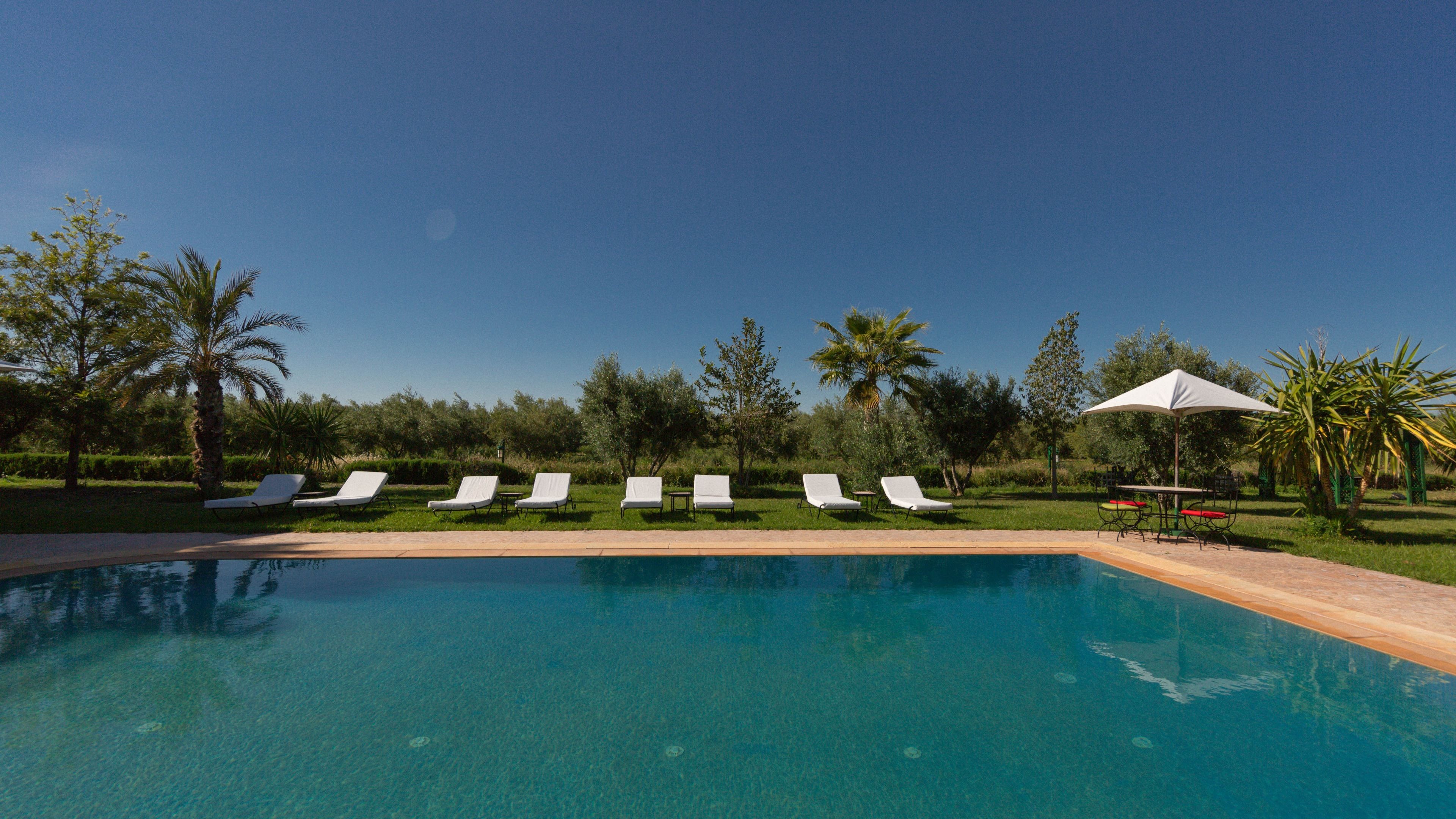Une piscine bordée de chaises longues blanches et agrémentée d’une table avec un parasol blanc.