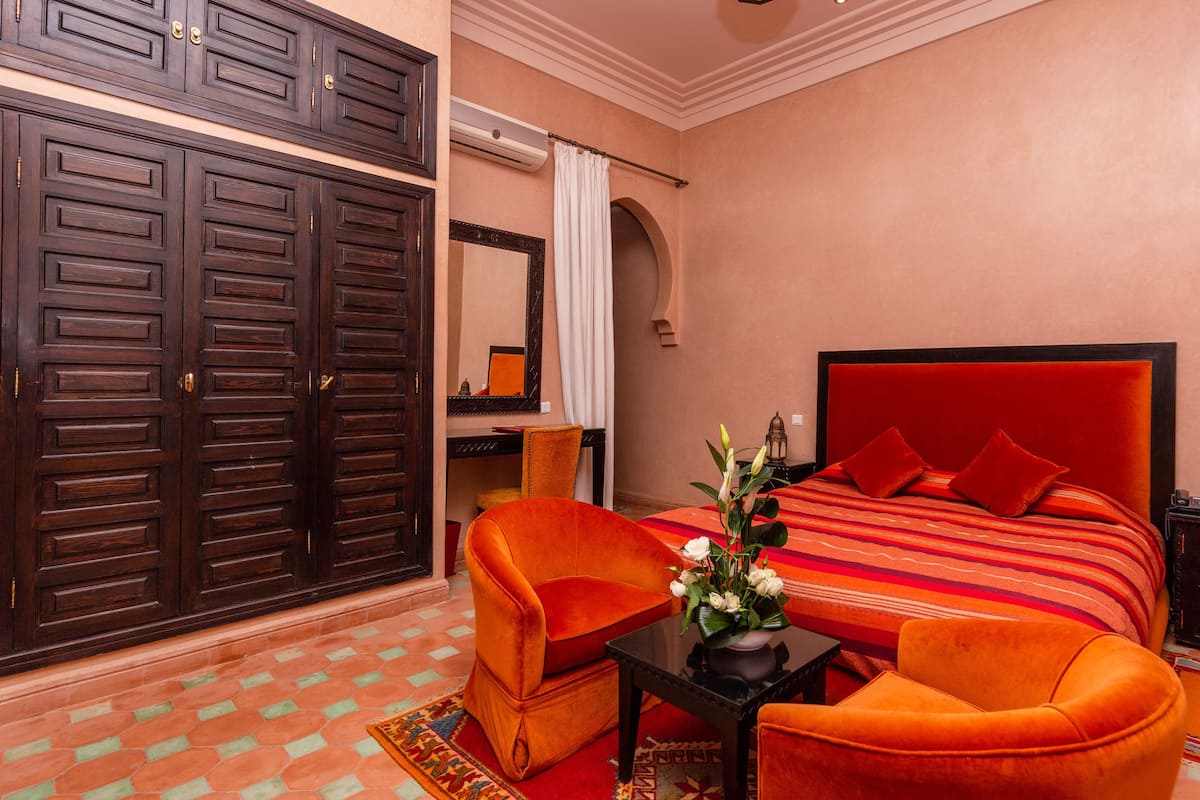 A hotel room with a large bed, two orange armchairs, a small table with a vase of flowers, a nightstand with a lamp, and a large wardrobe.