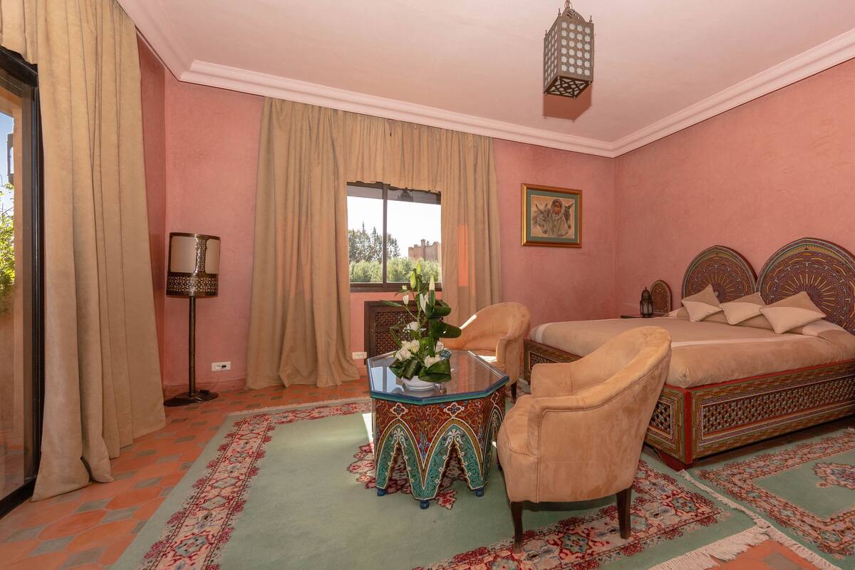 A bedroom with a bed, two chairs, a table with a vase, a nightstand, a lamp, and a window with curtains.
