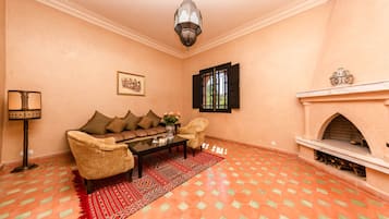 A living room with a sofa, armchairs, a coffee table, a patterned rug, a wall-mounted lamp, a framed picture, a window with black shutters, and a fireplace.
