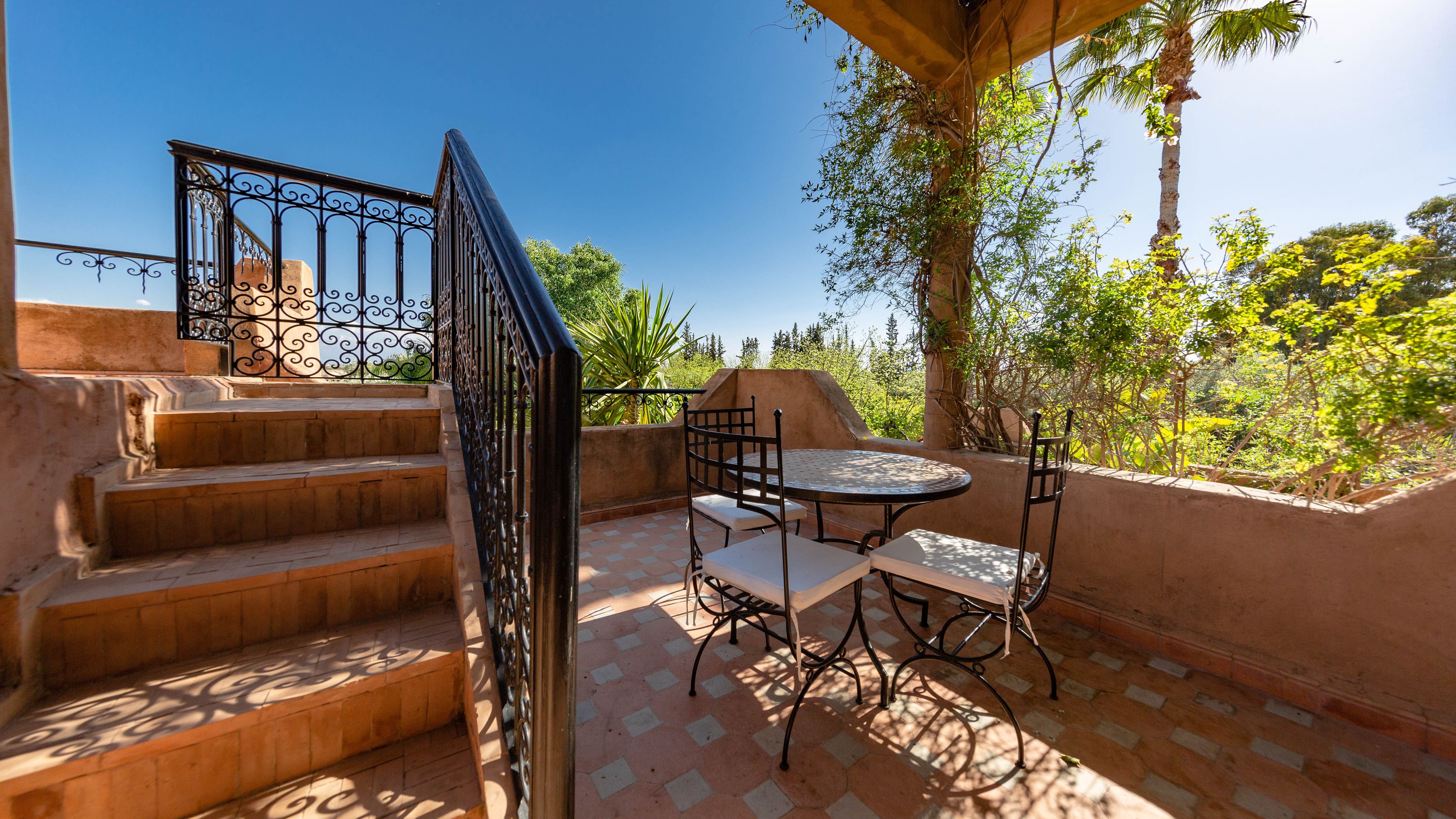 A patio area with a round table and chairs, a staircase with a metal railing, and a view of greenery and trees.