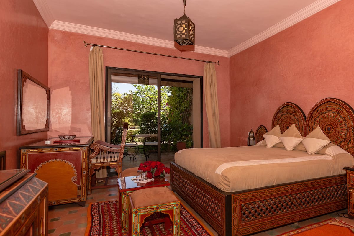A bedroom with a large bed, a wooden nightstand, a bedside table, a mirror, and a view of the outdoors through a sliding door.