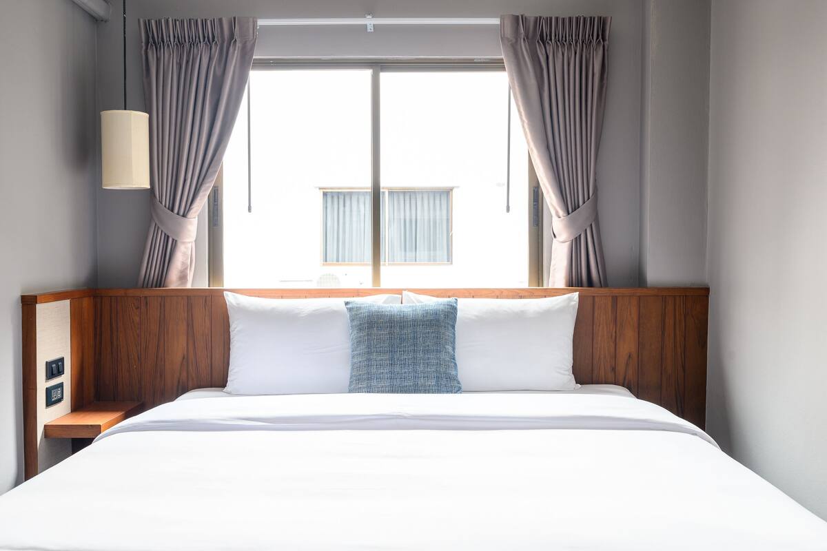 A neatly made bed with a wooden headboard, a bedside table with a lamp, and curtains on a window.