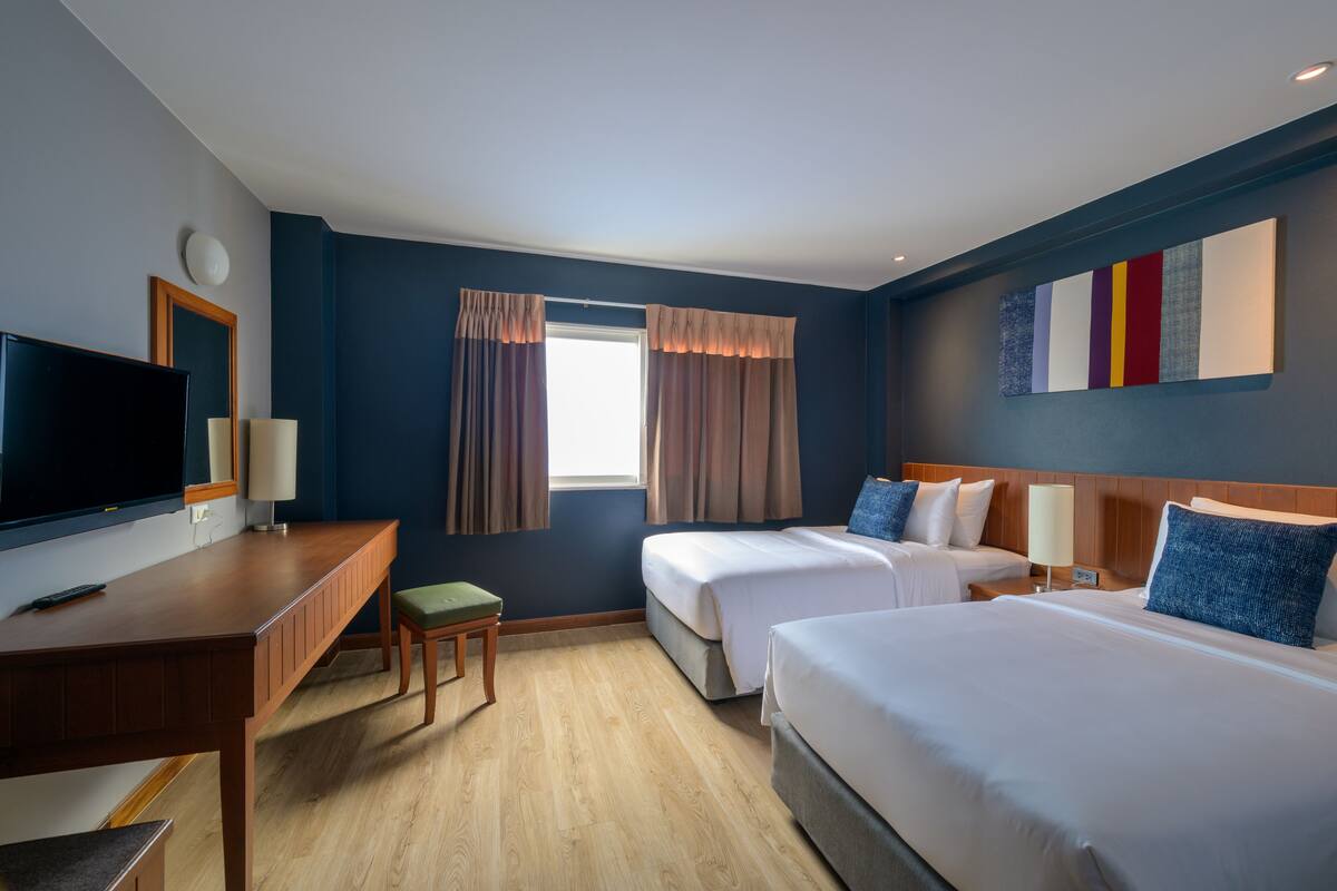 A hotel room with two beds, a desk, a TV, and a window with curtains.