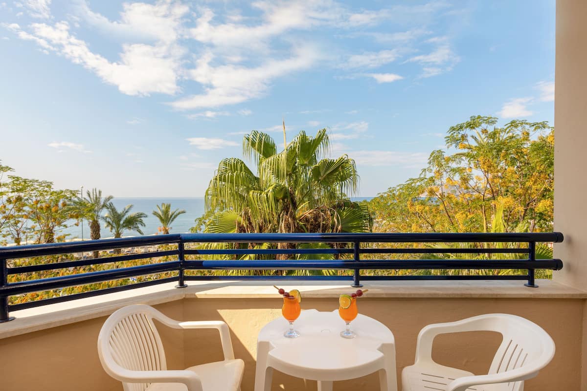 A balcony with two white plastic chairs, a small round table, and two drinks with garnishes. The balcony overlooks a view of palm trees and the ocean.