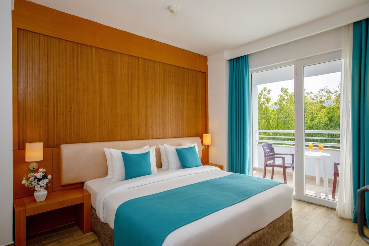 A hotel room with a large bed, wooden headboard, a nightstand with a lamp, a vase of flowers, and a balcony with a table and chairs.