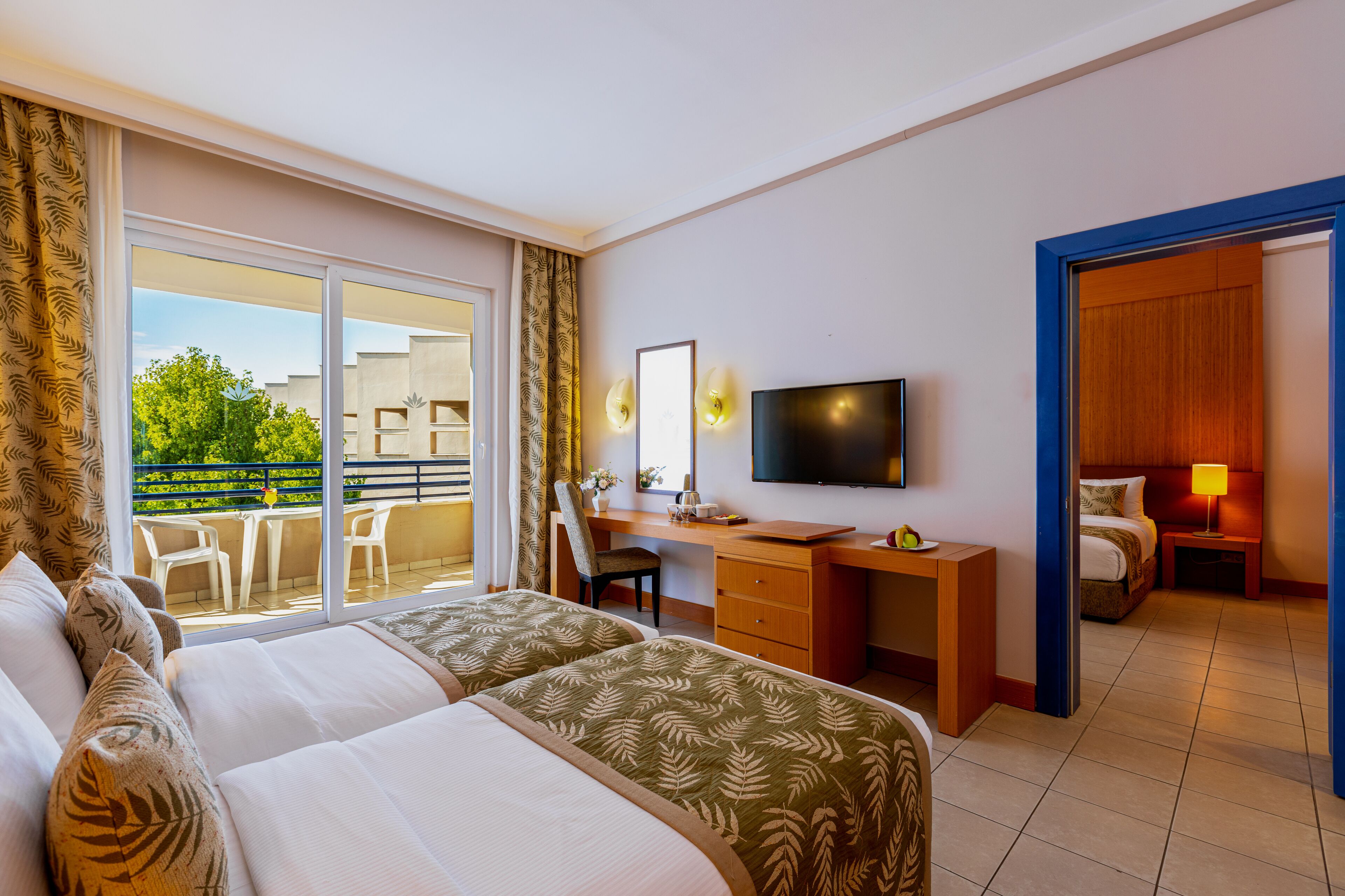 A hotel room with two beds, a desk, a TV, a balcony with chairs, and a view of greenery.