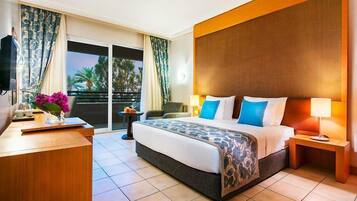 A hotel room with a large bed, a desk, a chair, a TV, and a balcony with curtains.