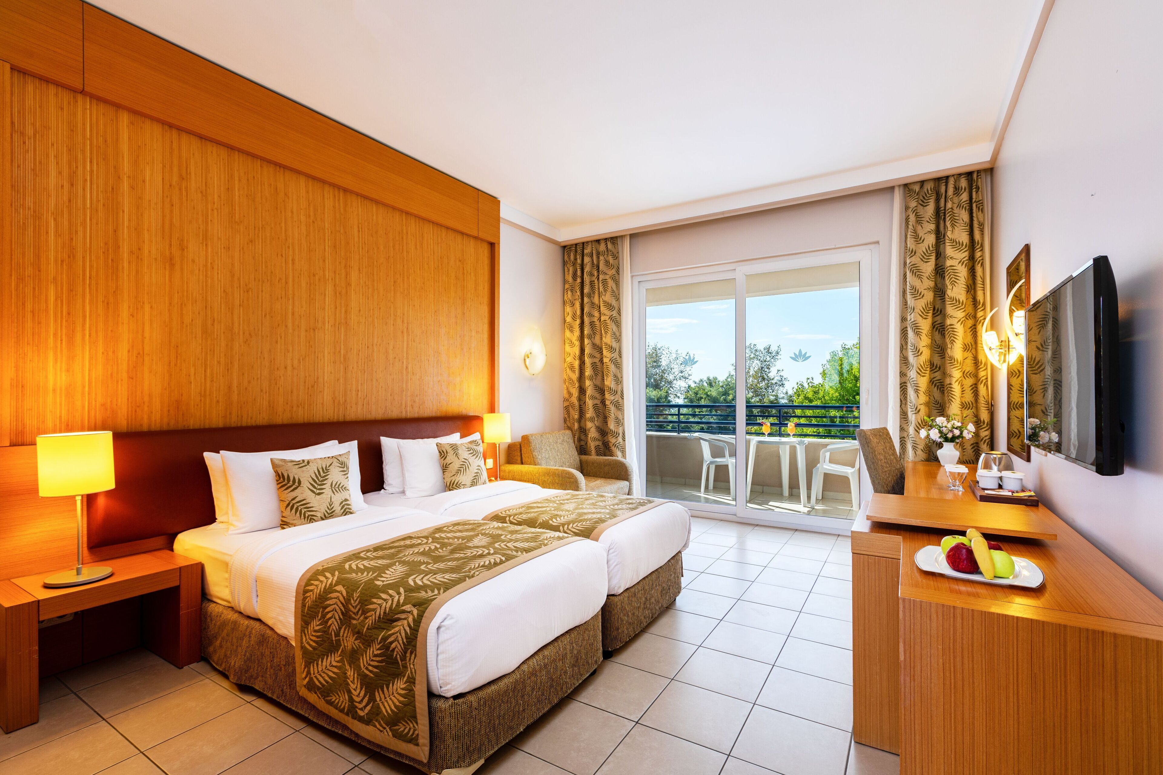 A hotel room with a large bed, a desk with a fruit plate, a TV, and a balcony with a view of greenery.