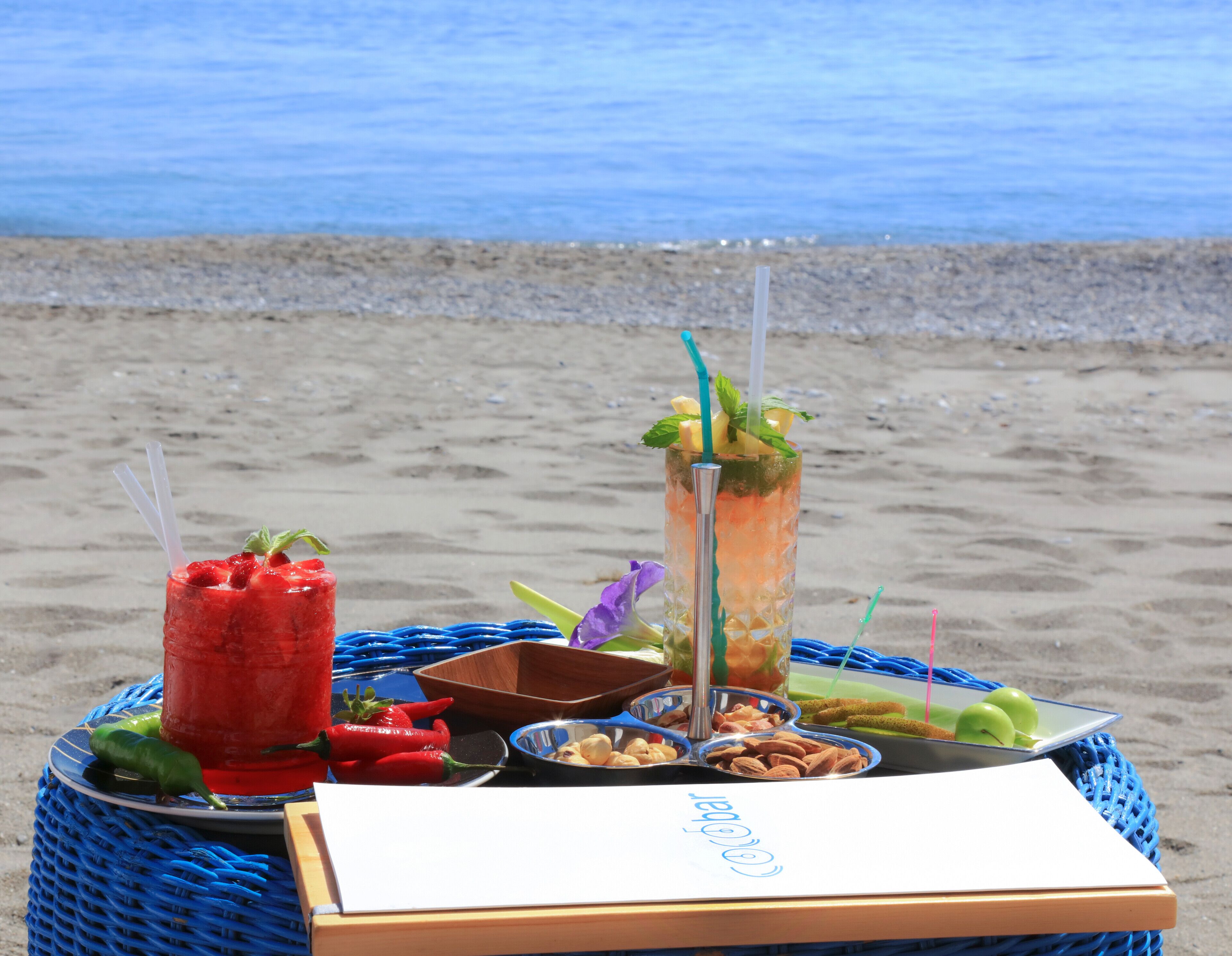 Ein Tisch am Strand mit einer Platte voller Speisen und Getränke, darunter ein Erdbeergetränk, ein tropischer Cocktail und verschiedene Snacks.