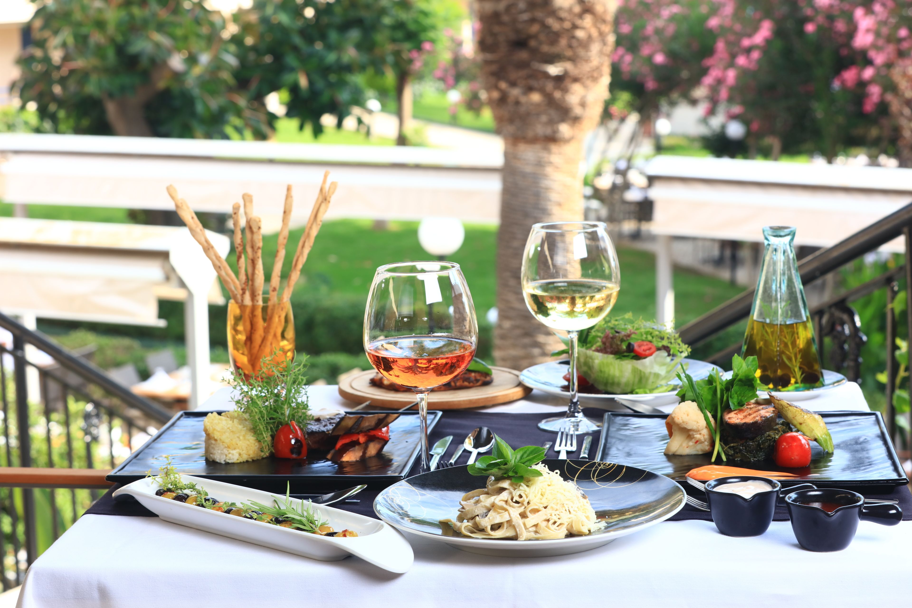 Mesa de comedor al aire libre con varios platos, copas de vino y una botella de aceite de oliva.