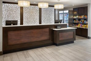Un espace d’accueil d’hôtel au design moderne, comprenant un mur décoré de motifs, un comptoir d’accueil et un mini-bar garni de collations.