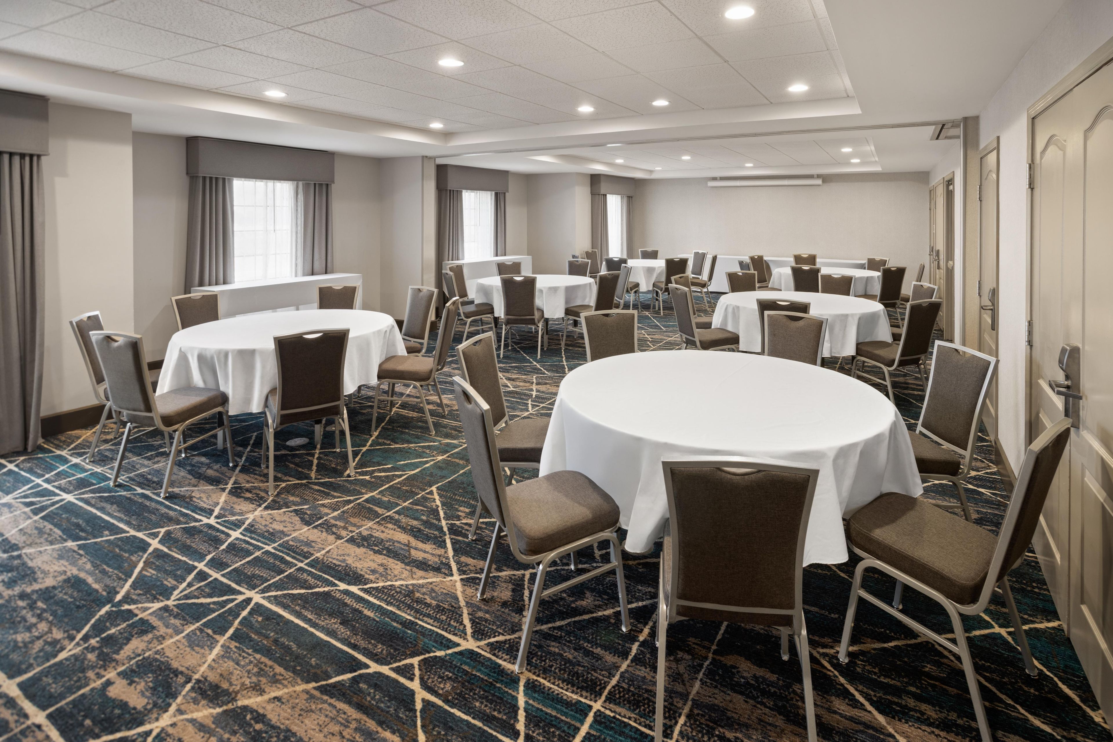 A hotel conference room with multiple round tables and chairs, carpeted flooring, and a ceiling with recessed lighting.