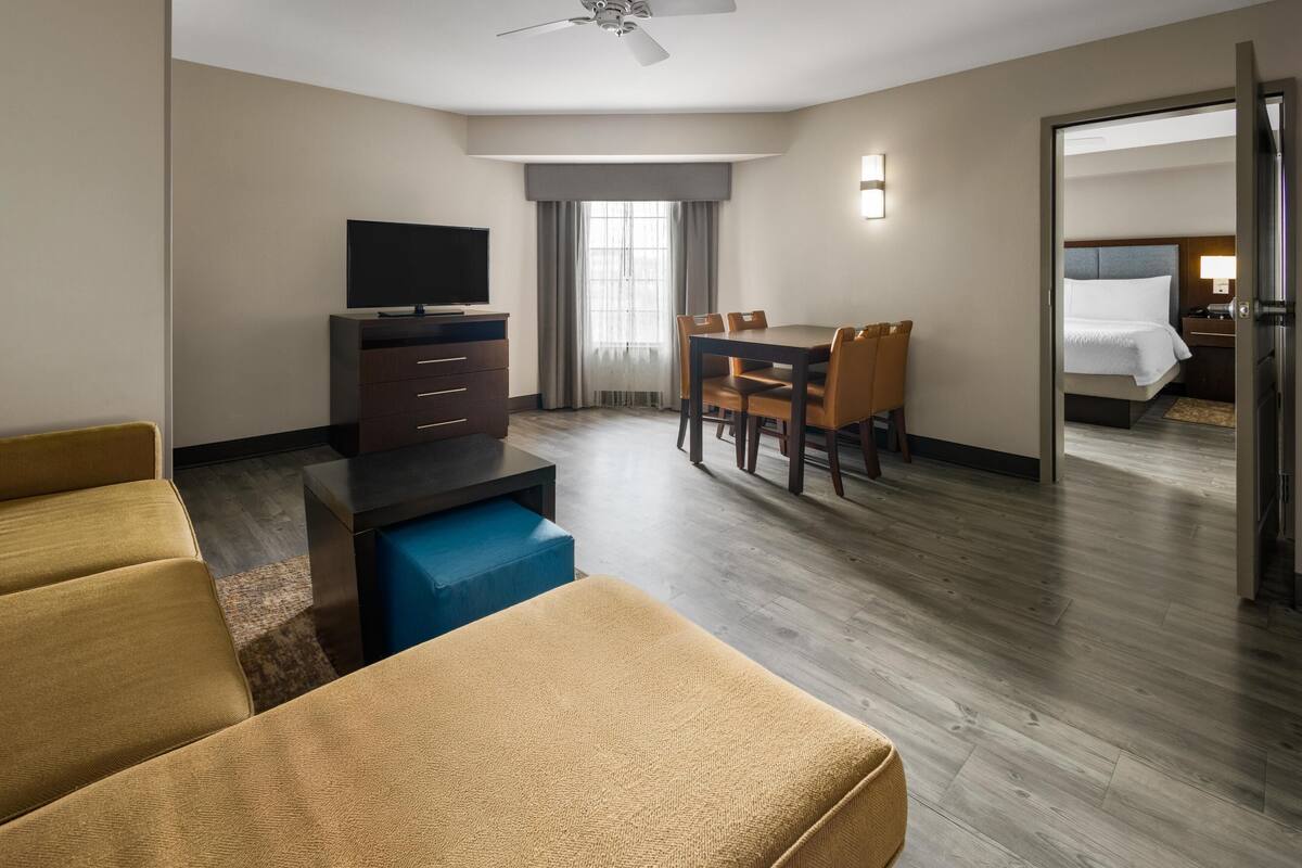 Une chambre d’hôtel équipée d’un canapé, d’un téléviseur posé sur un meuble, d’une table à manger avec des chaises, d’un lit et d’un ventilateur de plafond.