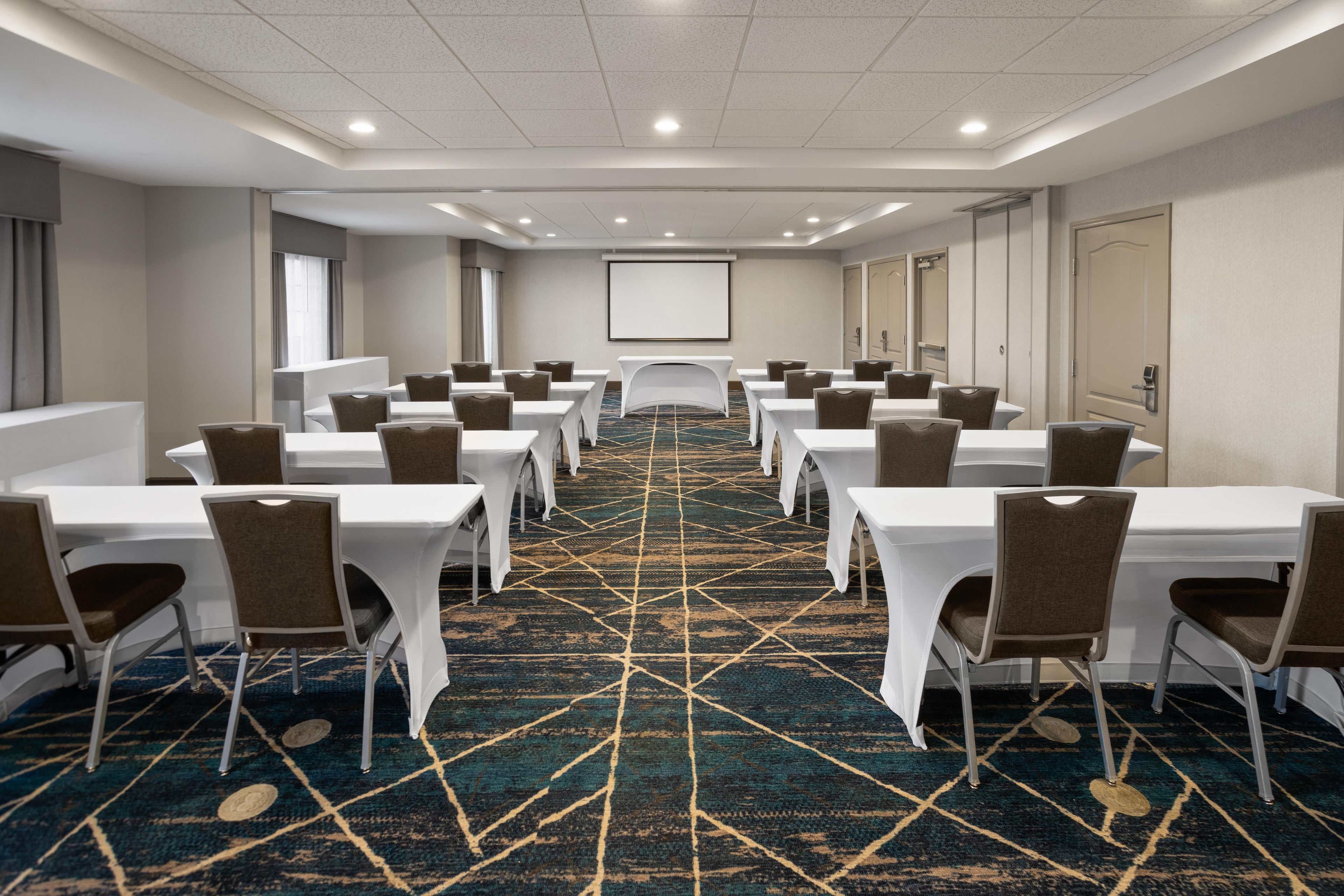 A conference room with rows of tables and chairs, a carpeted floor, and a projector screen on the wall.