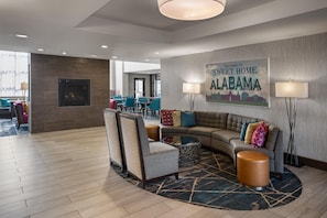 Een moderne hotellobby met een gebogen bank, fauteuils, een open haard en een bord met de tekst 'Welcome to Sweet Home Alabama'.