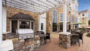 Een terras buiten met een barbecue van natuursteen, een zithoek en een pergola met lichtjes.