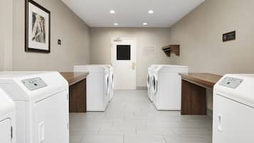 A laundry room with four white front-loading washers and dryers, wooden countertops, a framed artwork on the wall, and a sign indicating a fire exit.