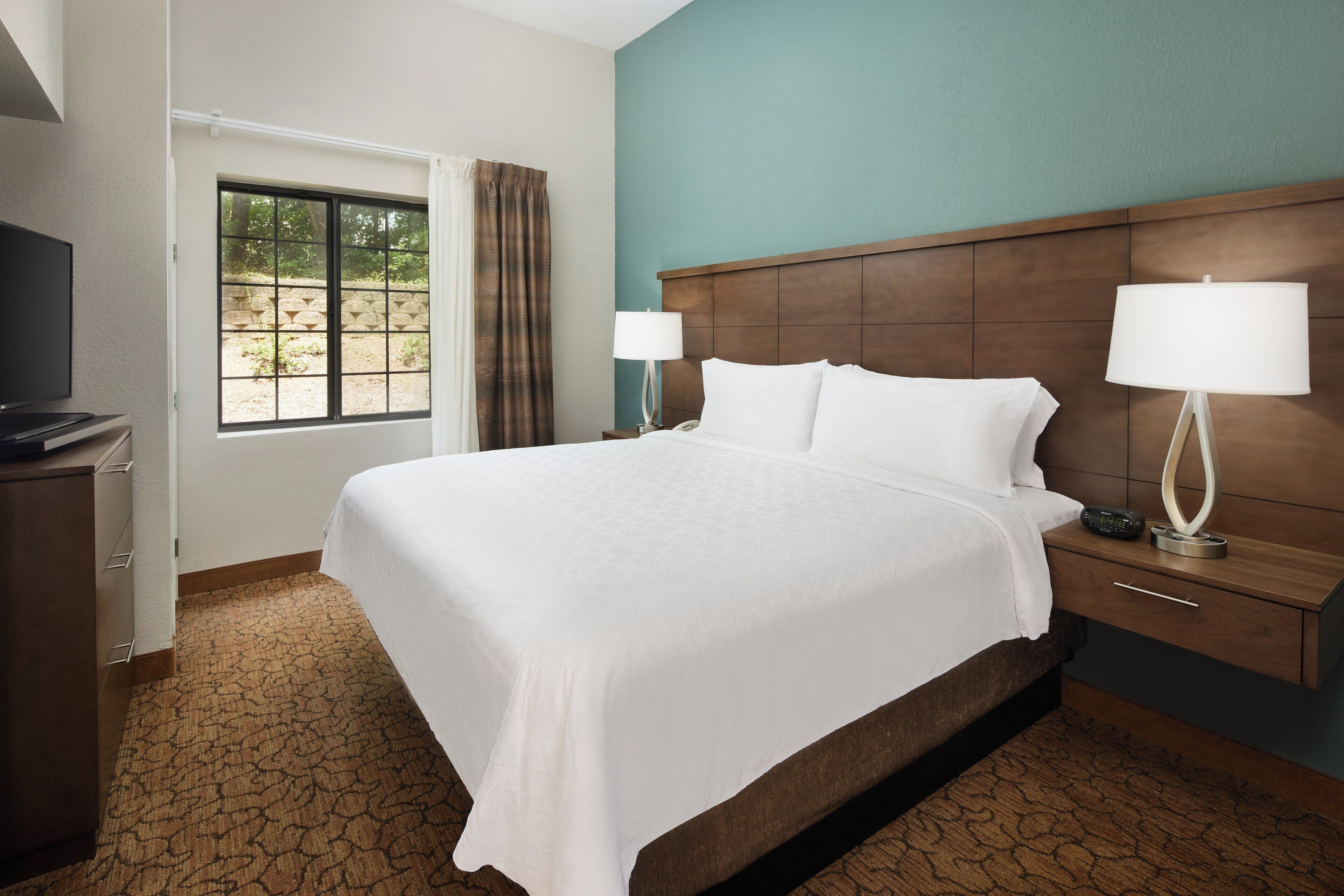 A hotel room with a large bed, a wooden headboard, a bedside table with a lamp, and a television on a stand.