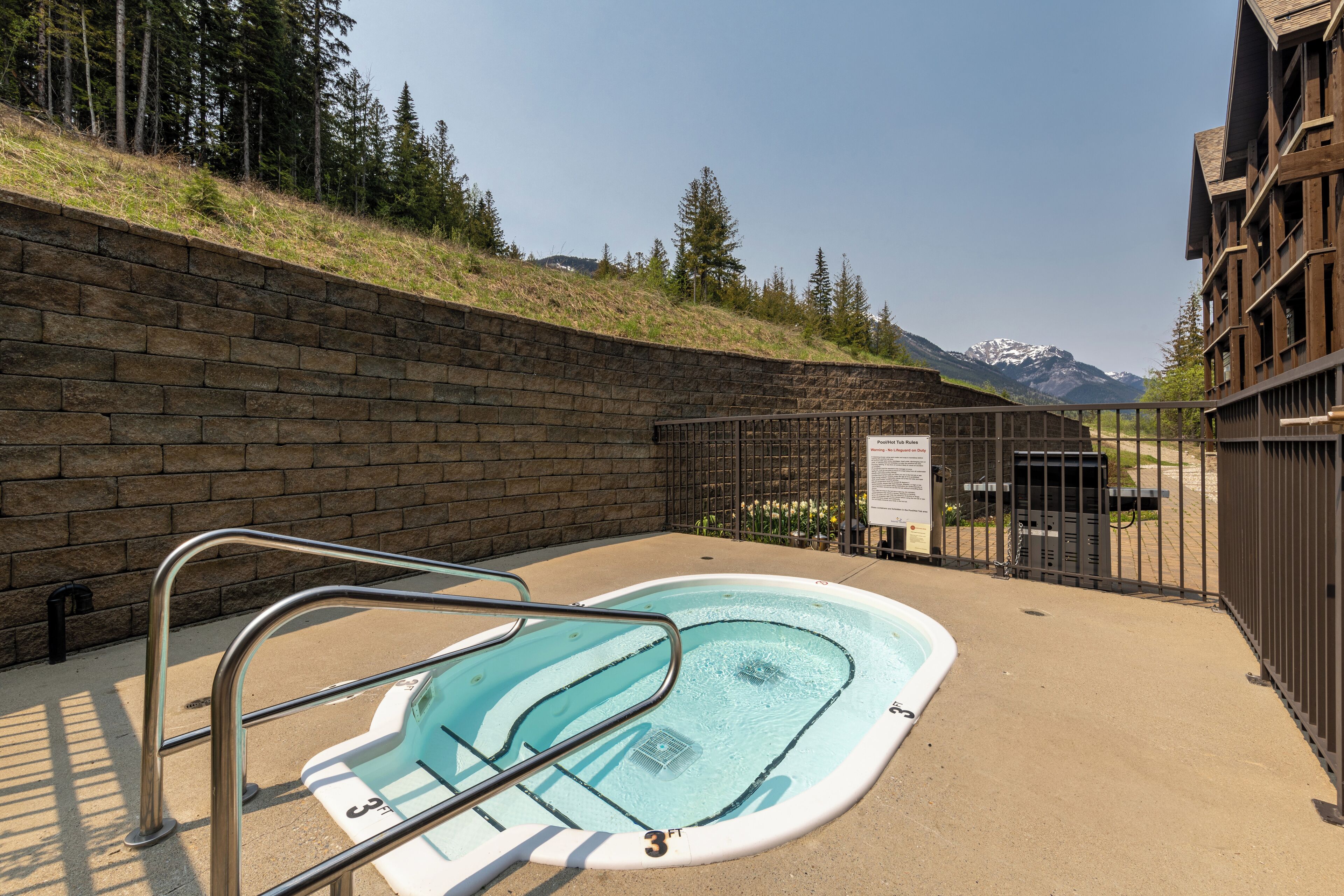 A small outdoor pool with a metal ladder, surrounded by a brick wall and a metal fence, with a mountain view in the background.