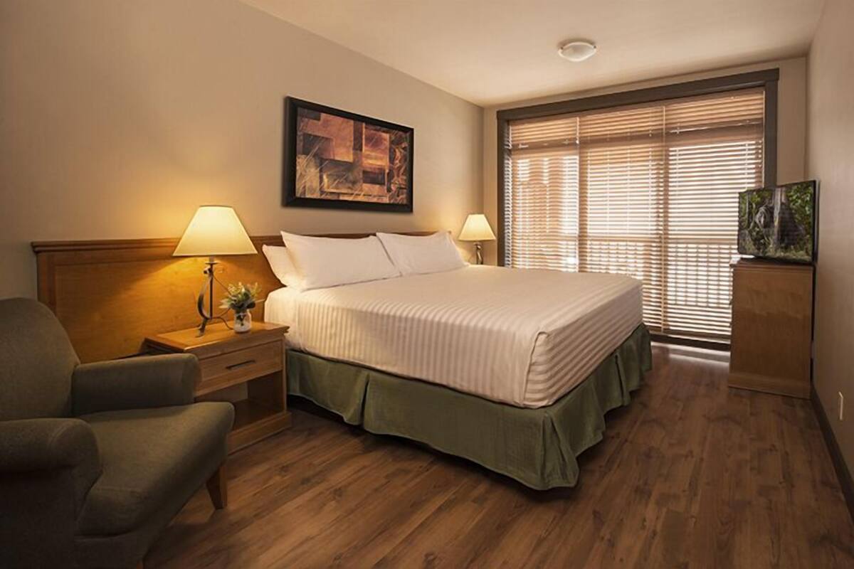 A hotel room with a large bed, two bedside lamps, a chair, a wooden headboard, a painting on the wall, and a window with blinds.