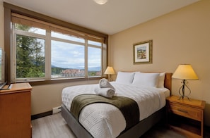A hotel room with a bed, two bedside lamps, a window with a view, and a painting on the wall.