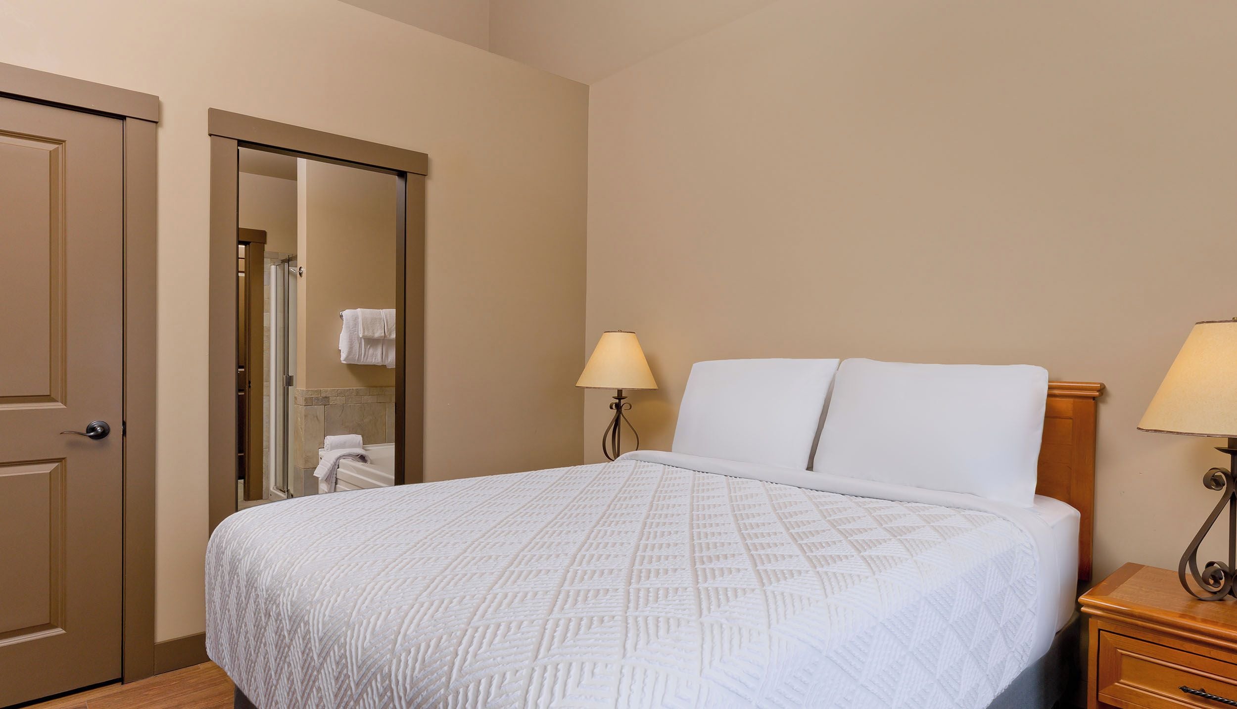 Ein Hotelzimmer mit einem Bett, zwei Nachttischlampen, einem Holznachttisch und einer Tür zu einem Badezimmer.