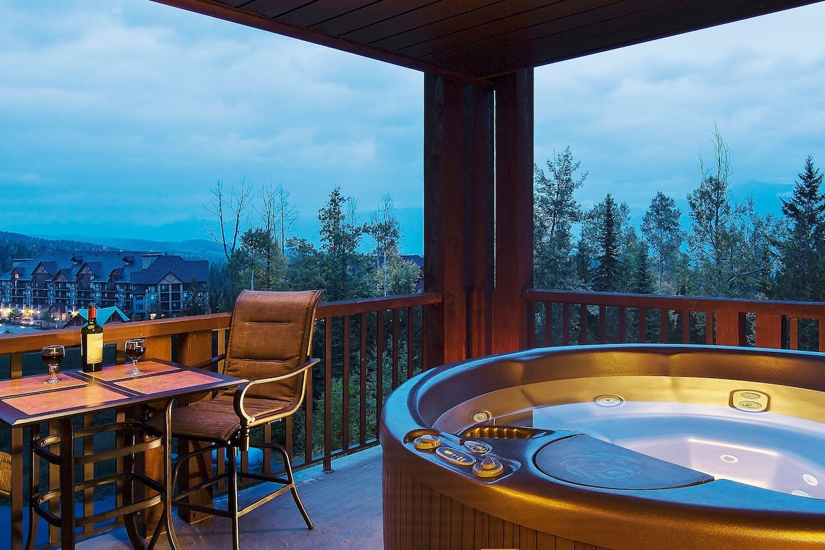 A wooden deck with a hot tub, a table with chairs, and a view of a mountain landscape.