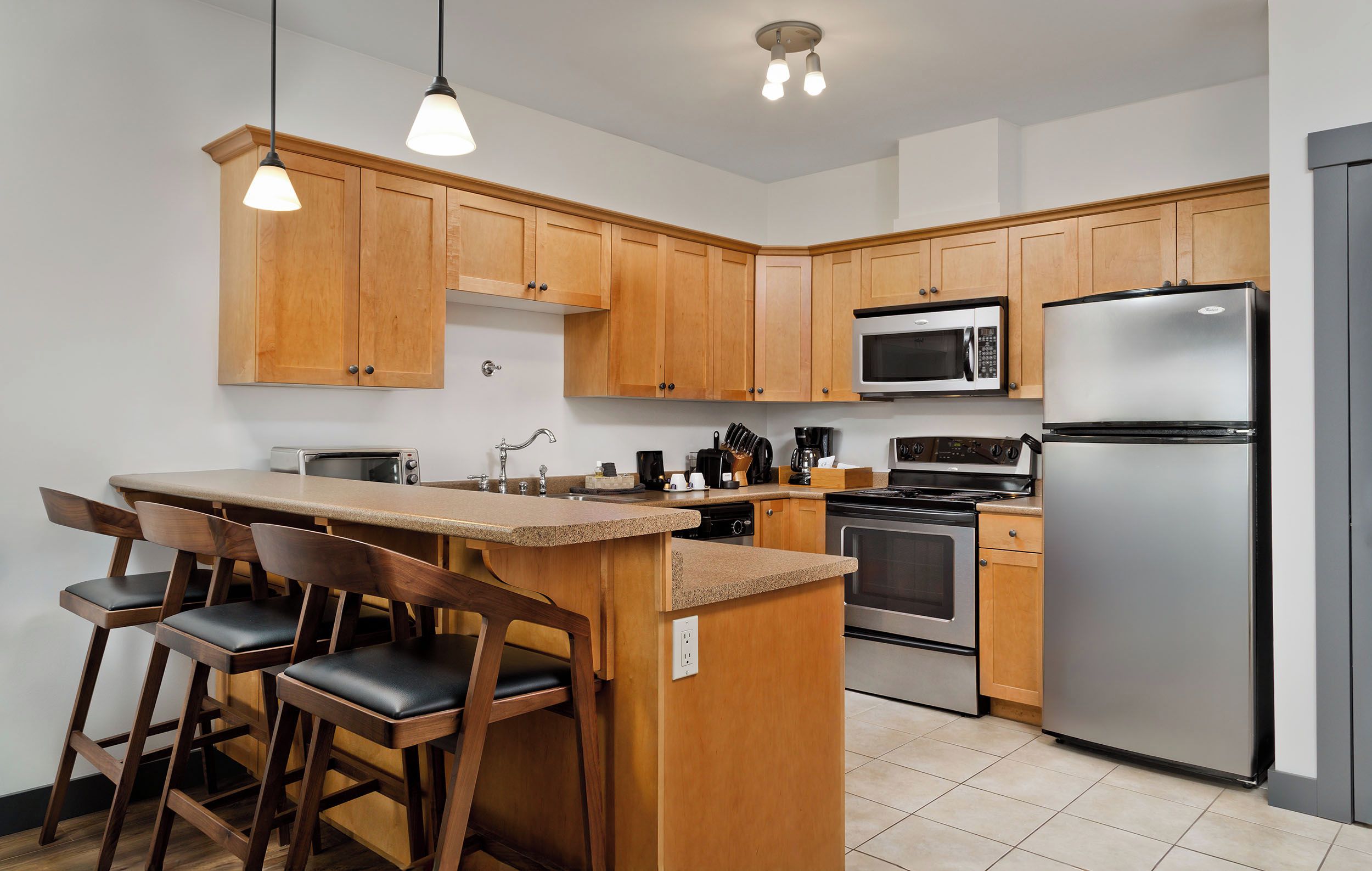 A modern kitchen with wooden cabinets, a stainless steel refrigerator, a microwave, a stove, and a bar area with stools.