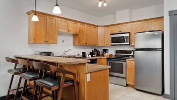 A modern kitchen with wooden cabinets, a stainless steel refrigerator, a microwave, a stove, and a bar area with stools.