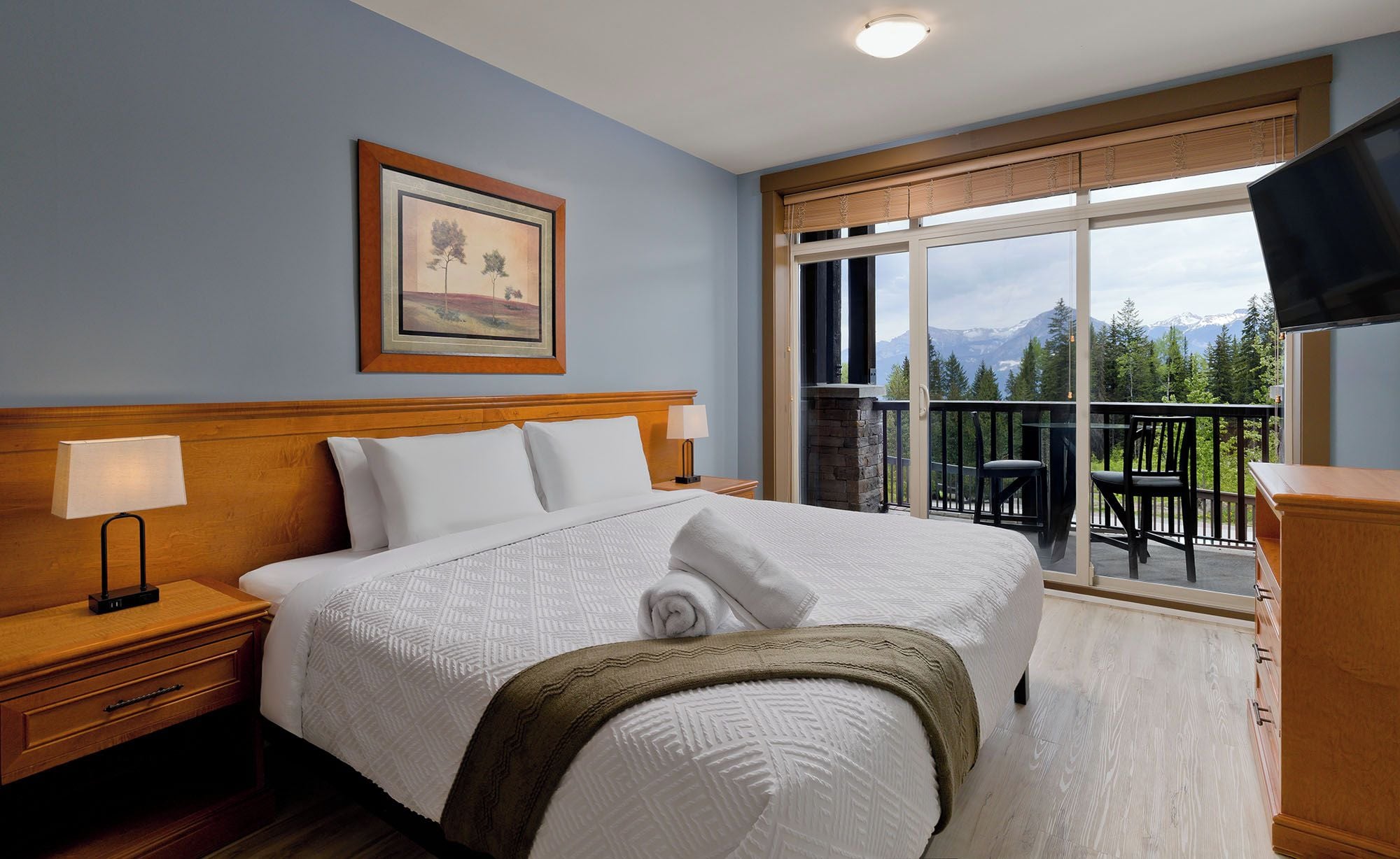 A hotel room with a large bed, a nightstand, a TV, a balcony with a view of mountains, and a framed picture on the wall.