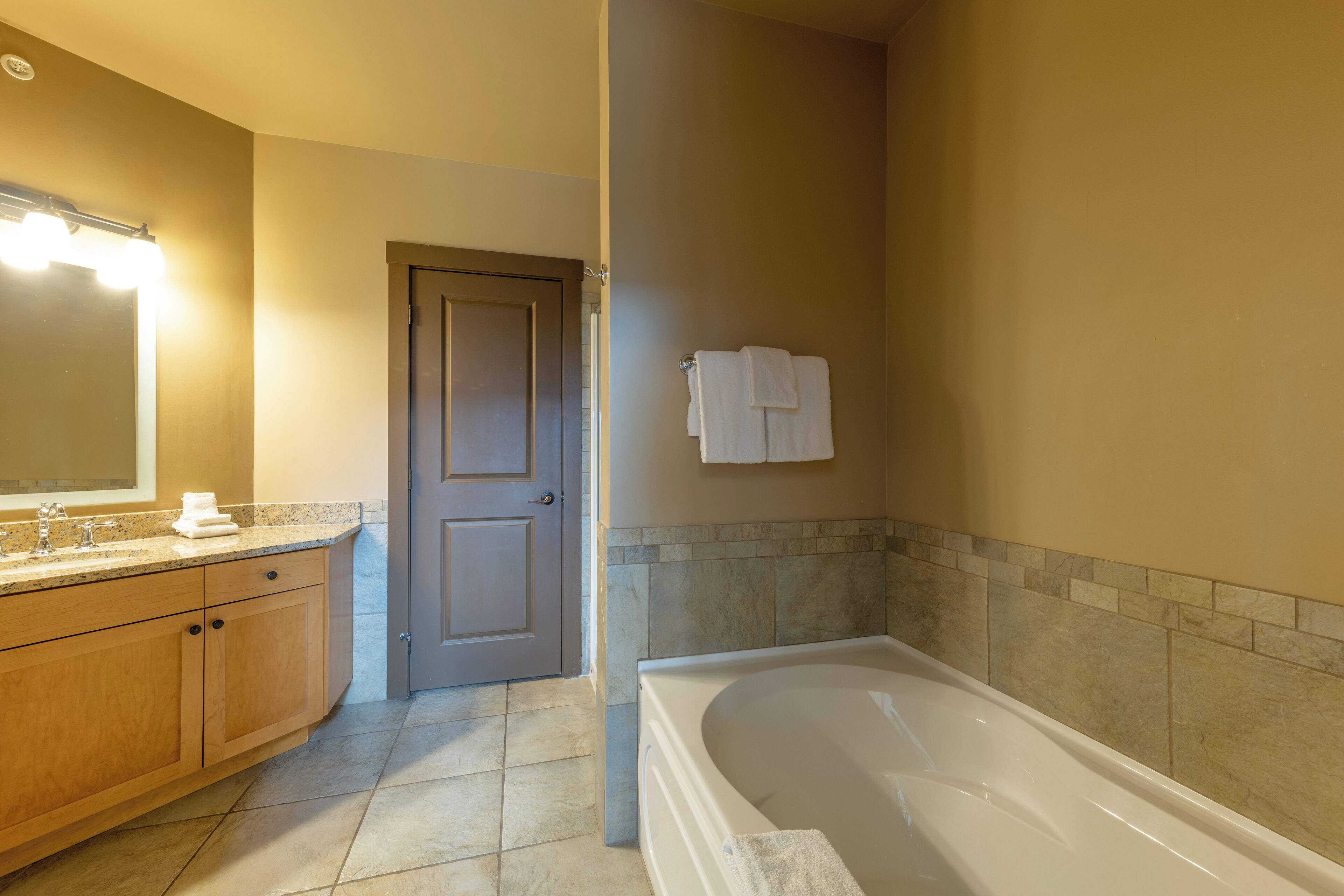 A bathroom with a bathtub, a vanity with a mirror and lights, and a door leading to another room.