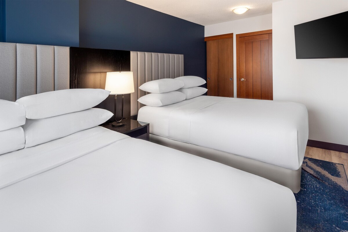 A hotel room with two beds, a nightstand with a lamp, a wooden wardrobe, and a television mounted on the wall.