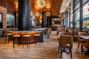 A modern bar area with high stools, a central bar counter, and a city view through large windows. - Embassy Suites Montréal by Hilton (Montreal)