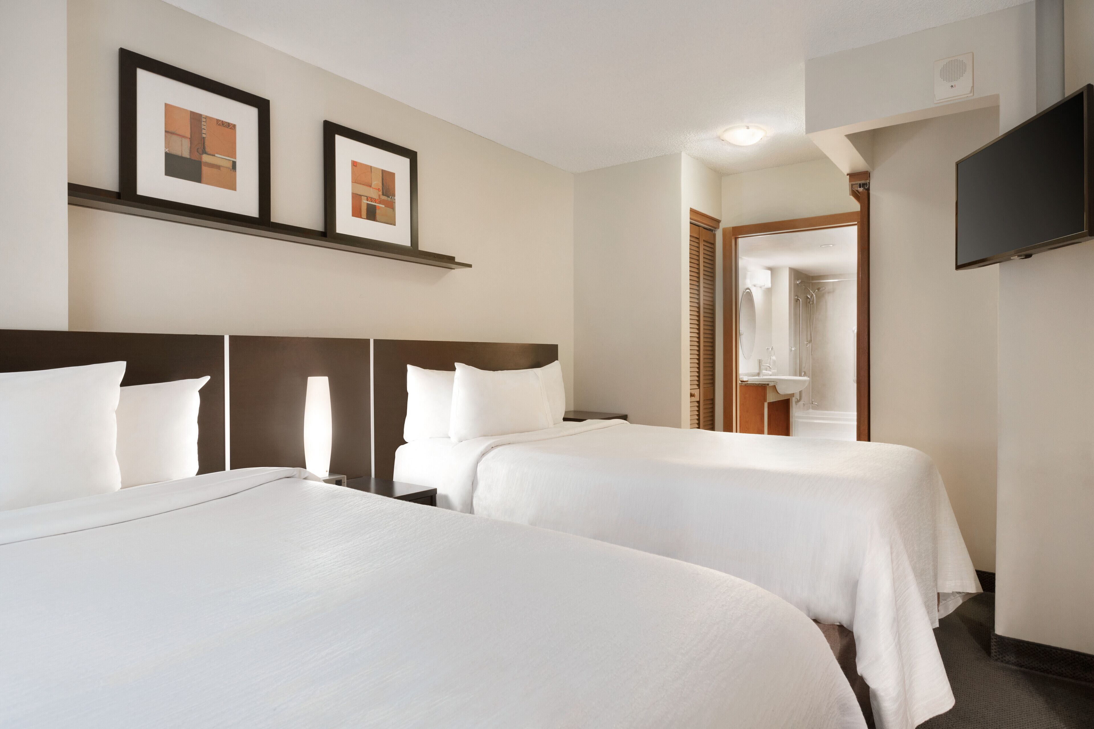 A hotel room with two beds, a TV, framed pictures on the wall, and a bathroom visible through an open door.