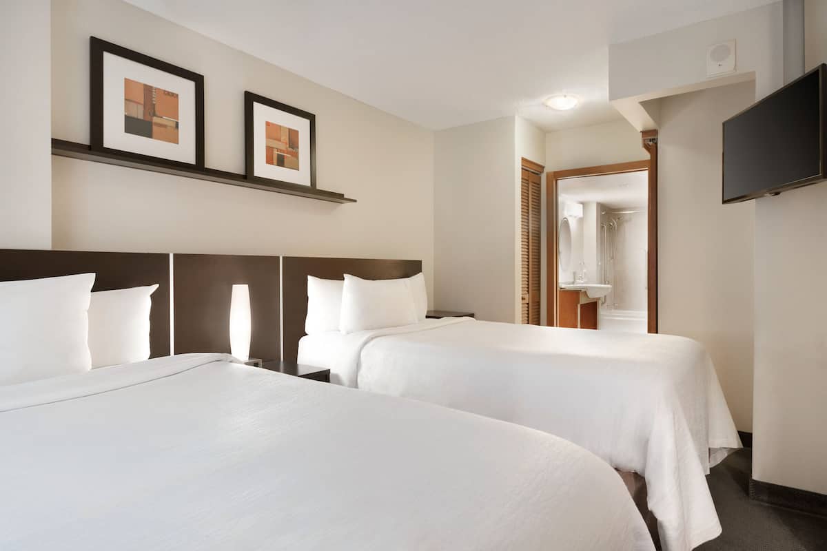 A hotel room with two beds, a TV, framed pictures on the wall, and a bathroom visible through an open door.