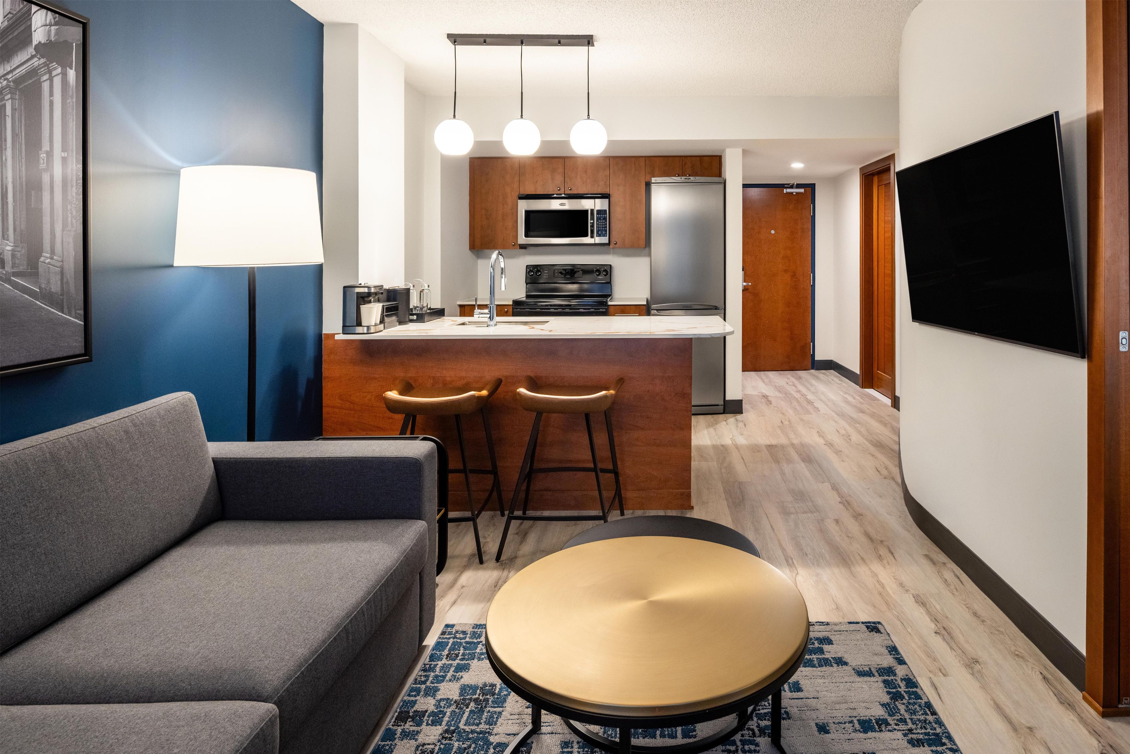 A modern hotel room with a sofa, a round table, a kitchen area with a microwave, a refrigerator, and a bar stool.