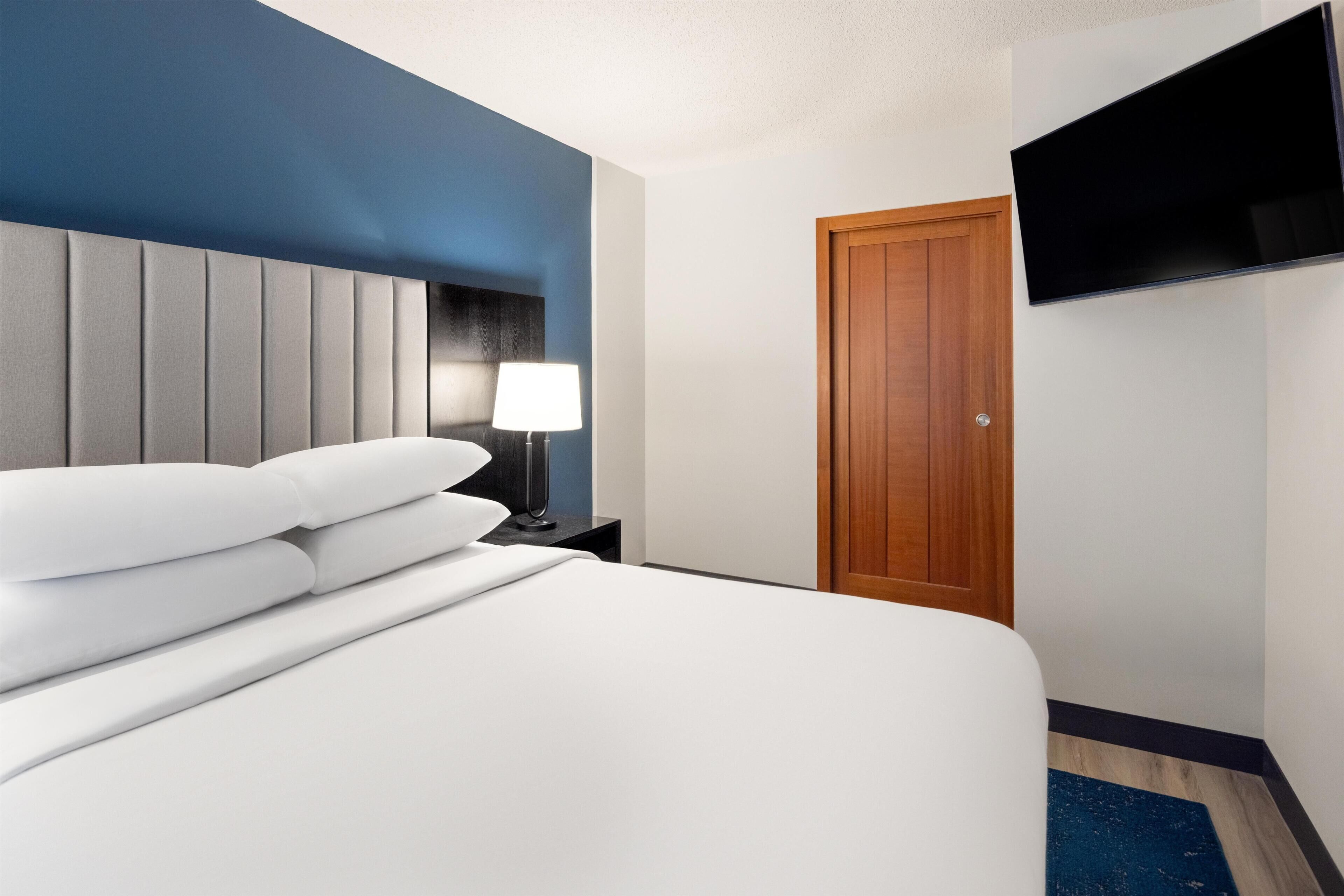 Habitación de hotel con cama, mesita de noche, lámpara, puerta de madera y televisor de pantalla plana montado en la pared.