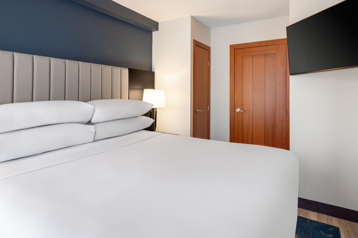 A hotel room with a bed, a nightstand with a lamp, a wooden door, and a flat-screen TV mounted on the wall.