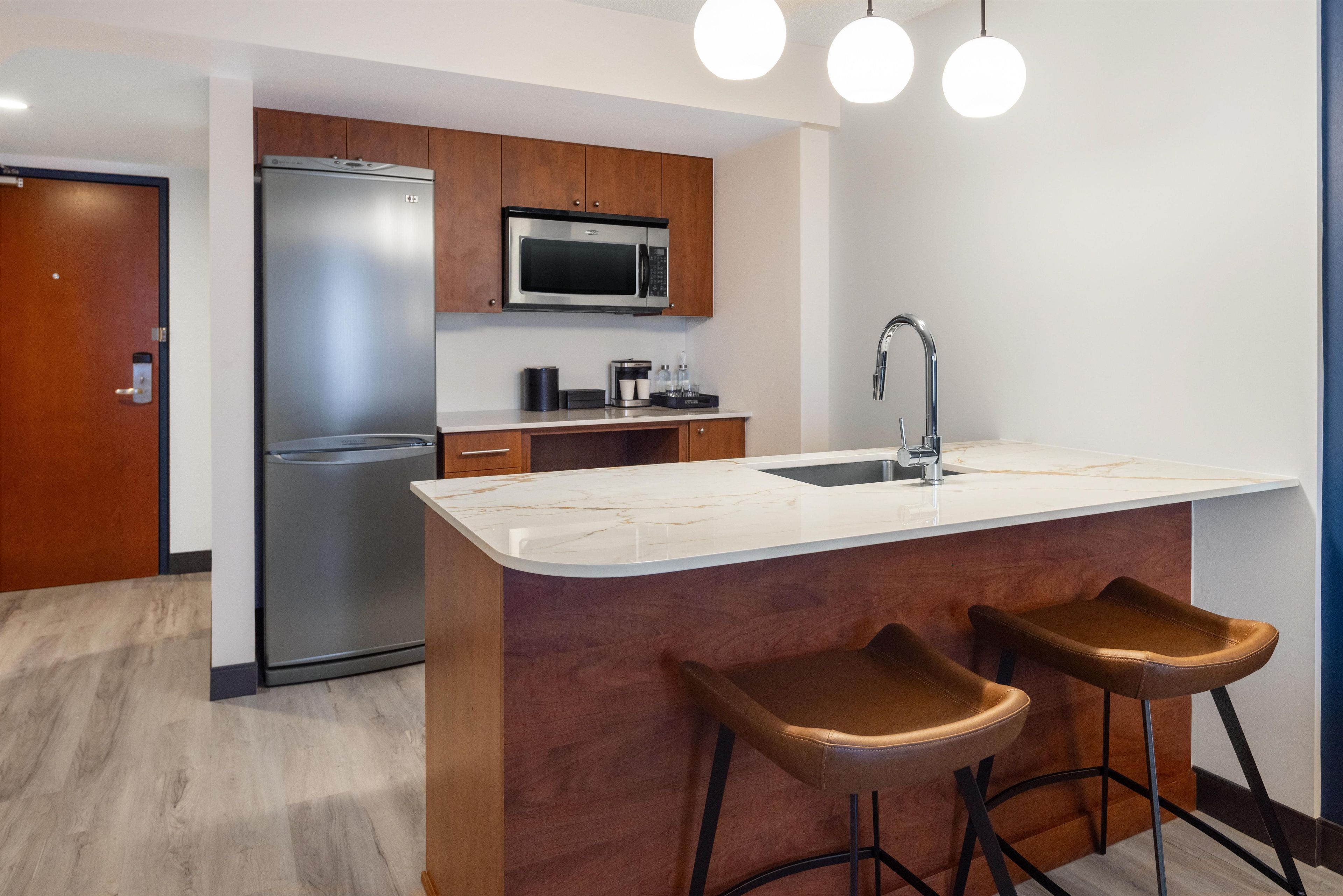 A modern kitchen with a built-in refrigerator, a microwave, a coffee maker, and a sink. There are two bar stools at the island.