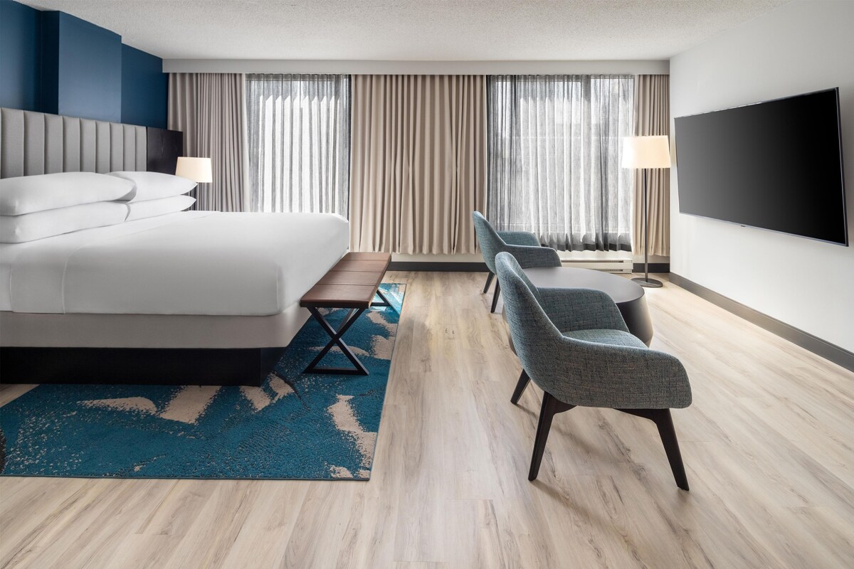 A hotel room with a bed, a desk, two chairs, a TV, and a large window with curtains.