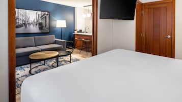 A hotel room with a bed, a sofa, a small round table, a bar area, and a television mounted on the wall.