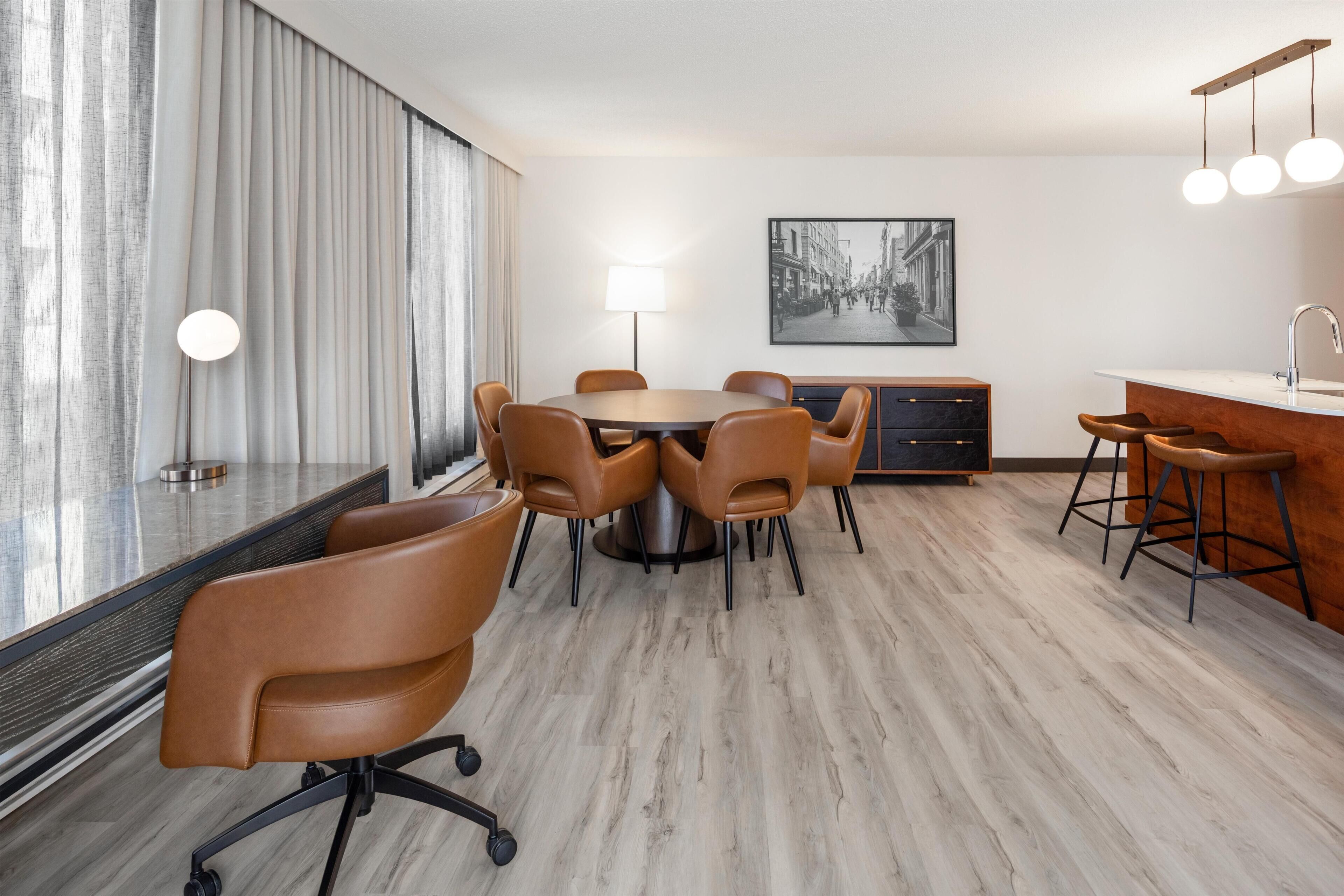 Una habitación de hotel moderna con zona de comedor, bar y un amplio ventanal con cortinas.