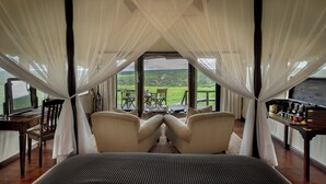 Quarto com uma cama grande, duas cadeiras, uma mesa pequena e vista para uma paisagem verde através de janelas amplas.